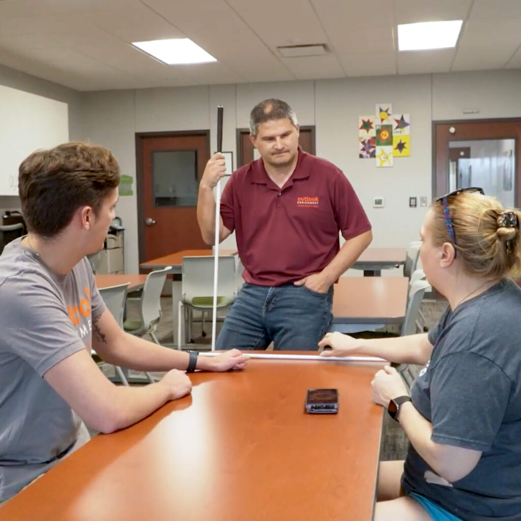 A man in a maroon shirt holds a cane while talking to a man and woman who sit at a table.